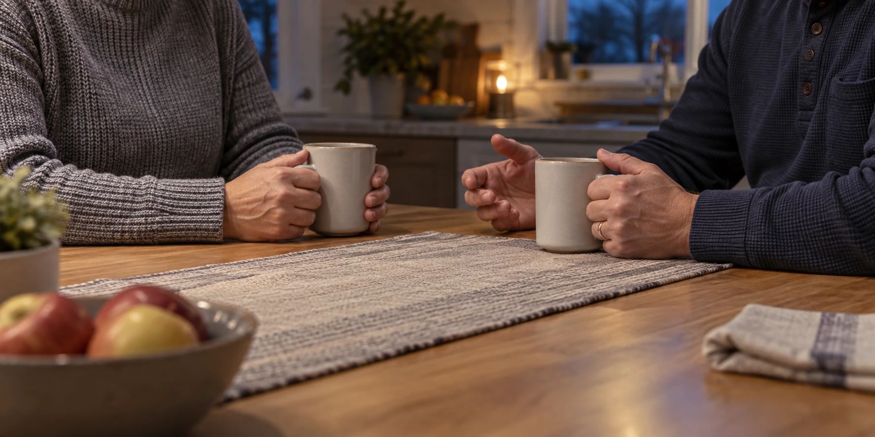 Two adults seated across a kitchen table with their hands near mugs.