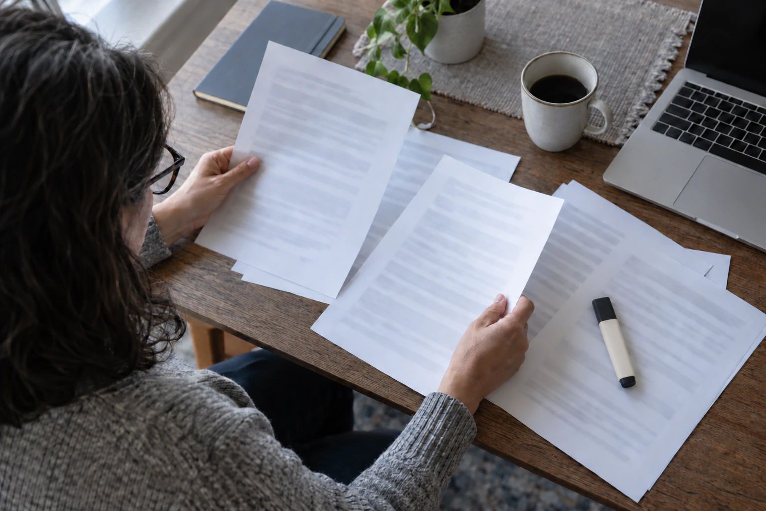 Person comparing sheets of paper at a desk beside a laptop and mug.
